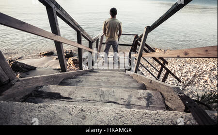 Junger Mann steht auf alte Holztreppe hinunter zur Küste Meeres. Vintage getönten Foto, alten Stil Fotofilter
