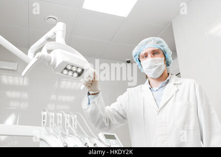 Ansicht von unten von einem Arzt Porträt Zahnarzt, Chirurg in Uniform und Maske im Gesicht, mit Brille, mit der Lampe in der Hand. Stockfoto
