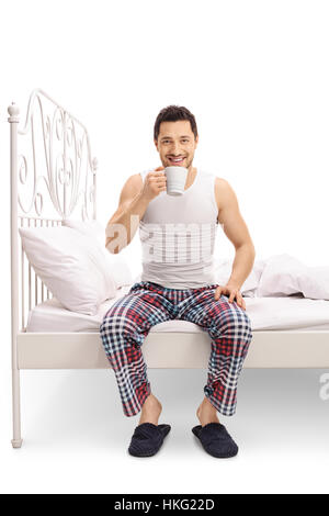 Junger Mann sitzt auf einem Bett und trinken aus einer Tasse isoliert auf weißem Hintergrund Stockfoto