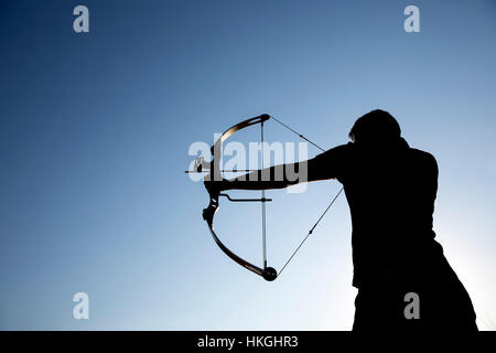 Eine Silhouette eines Bogenschützen zieht seine Verbundbogen und strebt nach oben mit klaren blauen Himmel als Hintergrund. Stockfoto