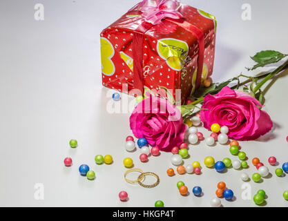 Leuchtend rosa Rosen mit verpacktes Geschenk-Artikel und Trauringe mit mehrfarbigen Papierkugeln in weißen Hintergrund isoliert. Stockfoto