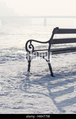 Bank im Schnee mit Blick auf zugefrorenen Fluss und Brücke an nebligen und sonnigen Tag. Winter-Stadtlandschaft. Stockfoto