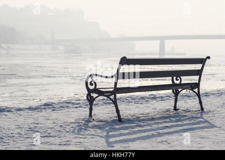 Bank im Schnee mit Blick auf zugefrorenen Fluss und Brücke an nebligen und sonnigen Tag. Winter-Stadtlandschaft. Stockfoto