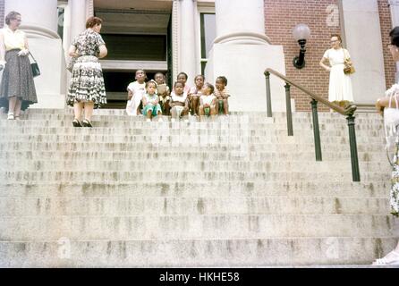 Vier reife kaukasische Frauen, die Kleider und Fersen tragen, stehen an verschiedenen Positionen auf der Marmortreppe des Maryland State House und schauen mit amüsierter Munterung auf eine kleine Gruppe von jungen afroamerikanischen Mädchen, die Kleider tragen und in zwei Reihen auf den Stufen des Gebäudes sitzen, Originaltitel lautet: "Farbige Kinder, Annapolis Capitol", Annapolis, Maryland, 1952. Stockfoto