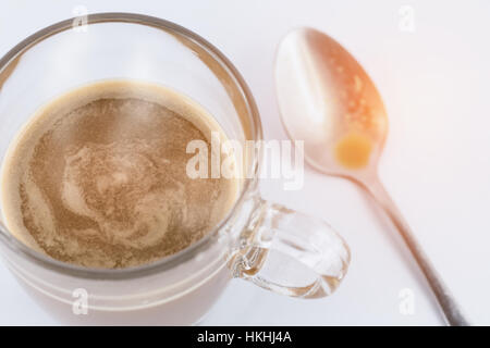 Enger Fokus auf heißen Kaffee in Klarglas-Cup am weißen Tisch. Verschwommen Teelöffel und orange warmes Sonnenlicht als Hintergrund. Stockfoto