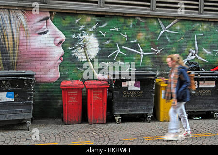 Graffiti in Glasgows Hinterstraße, blonde Frau bläst einen Löwenzahn in Windturbinen, erzeugt grüne Energie, in der Nähe von Mülltonnen Stockfoto