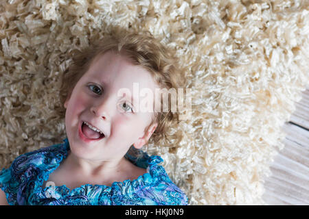 Ein junges Mädchen mit blonden Locken legt auf den Boden auf einem weichen Teppich spielerisch in die Kamera schauen und lachen; St. Albert, Alberta, Kanada Stockfoto