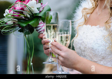 Eine Braut hält zwei Gläser Champagner und einen Blumenstrauß. Stockfoto