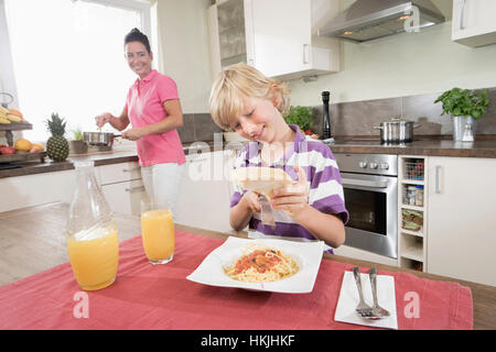 Junge Käse reiben, über Spaghetti, während seine Mutter kochen Essen, Bayern, Deutschland Stockfoto