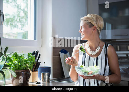 Junge Frau in der Küche Salat essen und lächelnd, Bayern, Deutschland Stockfoto