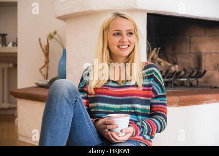 Schöne junge Frau Tasse Tee und sitzen auf dem Boden vor dem Kamin im Wohnzimmer, Bayern, Deutschland Stockfoto
