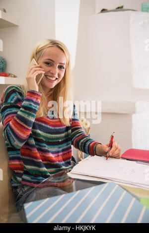 Junge Studentin Hausaufgaben und telefonieren mit Handy in die Wohnzimmer, Bayern, Deutschland Stockfoto