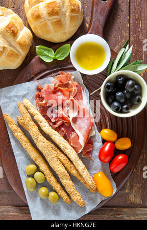 Prosciutto Crudo mit Brot, Oliven, Tomaten, Brot-Sticks, Olivenöl und Basilikum auf alten Holztisch. Stockfoto