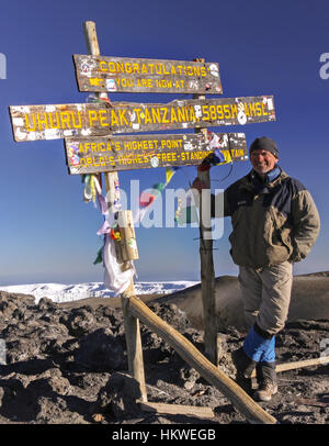 Männlicher Wanderer Im Stehen Vertikales Hochformat, Kilimandscharo Summit Top. Uhuru Peak, Tansania mit ikonischem Holzpfosten-Schild und Hintergrund des blauen Himmels Stockfoto