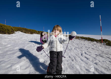 Blonde Kind drei Jahre alt mit Schneeball in der hand bereit, neben Frau Mutter, mit weißen und lila Mäntel auf Schnee im Winter Mountai werfen suchen Stockfoto