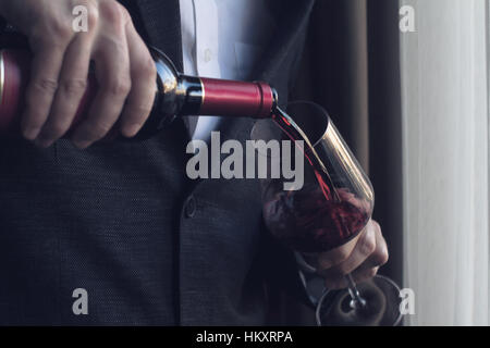 Horizontale Nahaufnahme von kaukasischen Mann im schwarzen Anzug und weißem Hemd strömenden Rotwein in ein hohes Glas in einer bar Stockfoto