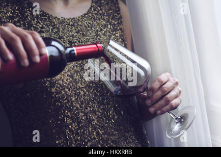 Horizontal in der Nähe der kaukasischen Retro-Frau in gold Pailletten Vintage verkleiden gießen sich Rotwein in ein großes Glas zu trinken in der Nähe eines Fensters Stockfoto