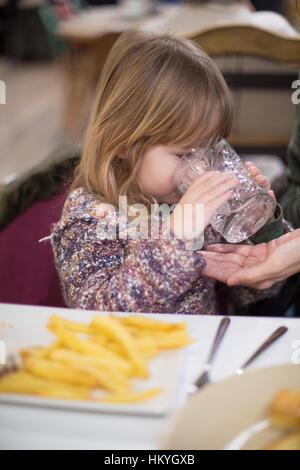 Drei Jahre blonde süße kaukasische Kind mit Strickpullover Trinkwasser aus Kristallglas, mit Pommes frites weißen Teller im restaurant Stockfoto