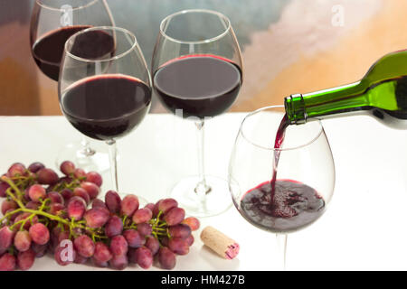 Foto von Rotwein in Glas Flasche mit mehr gegossen verschwommen volle Weingläser im Hintergrund, und auch aus Fokus Trauben und Kork Stockfoto