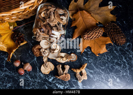 Trockene weiße Pilze im Glas und auf den Tisch. Gelb aus Holz Korb am hinteren Hintergrund, trocknen mehrere Eichenlaub, Eicheln und Tannenzapfen. Stockfoto