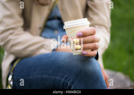 Ein junges Mädchen hält in ihrer linken Hand mit einem hellen Maniküre Einweg-Kaffeetasse.  Im Hintergrund sehen Sie einen grünen Park. Das Mädchen in jeans Stockfoto