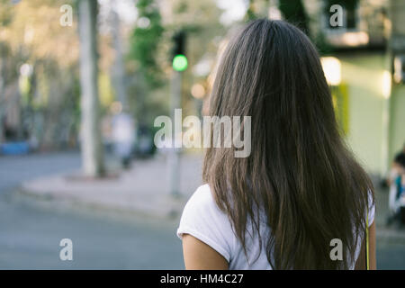 Junge weibliche Fußgänger überqueren Sie die Straße auf eine grüne Ampel. Das Mädchen hat lange Haare, trägt sie ein weißes T-shirt. Hintere Ansicht Nahaufnahme. Frau in Stockfoto