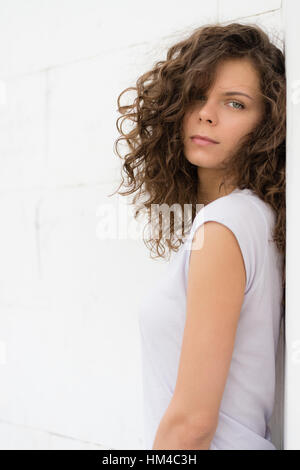 Junge, attraktive Frau mit lockigen braunen Haaren auf einem weißen Hintergrund. Porträt eines schönen Mädchens in Tageslicht im Freien. Stockfoto