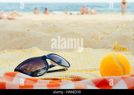 Sonnenbrillen und ein Tennisball liegen auf einer Decke auf dem Hintergrund des Strandes im Sommer Stockfoto