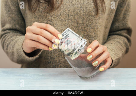 Mädchen im braunen Pullover mit gelben Maniküre bringt Geld in ein Glas, Nahaufnahme Stockfoto