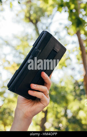 Weibliche Hand hält ein e-Book in der Abdeckung auf dem Hintergrund der Bäume Stockfoto