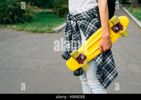 Mädchen in Jeans und ein kariertes Hemd ist mit gelben Kunststoff-Skateboard in Händen im Park spazieren. Stockfoto