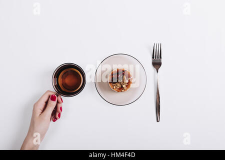Tasse Tee, Kuchen und Gabel auf einem weißen Tisch, eine weibliche Hand mit roten Maniküre hält eine Tasse, Ansicht von oben Stockfoto