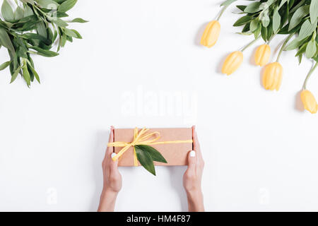 Weibliche Hände halten Geschenkbox mit gelben Band auf dem Tisch in der Nähe von Tulpen, Ansicht von oben Stockfoto