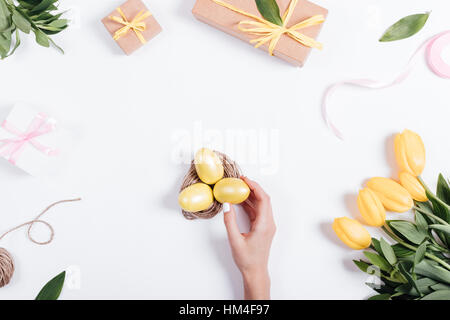 Frauen Hand legt gelbe Ostereier in einem Nest auf einem Tisch in der Nähe von Tulpen, Ansicht von oben Stockfoto