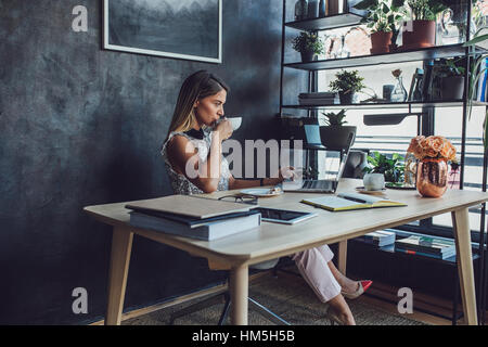 Geschäftsfrau trinkt Kaffee während der Arbeit am Laptop im Heimbüro Stockfoto