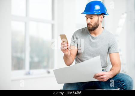 Gut aussehend Mechaniker oder Baumeister in Zusammenarbeit mit Zeichnungen und Telefon mit der Renovierung der Wohnung innen Helm Stockfoto