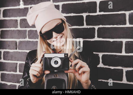 Porträt der hübsche blonde Haare drausen junge Teenager Hipster Woman Modell mit Retro-Foto-Kamera trägt einen rosa Hut, schwarze Lederjacke. Fashion-Look. Stockfoto