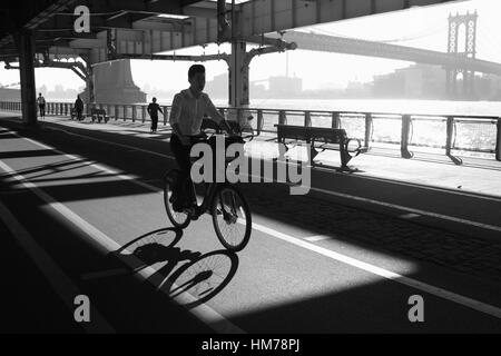 Mann, Radfahren entlang der East River Esplanade unter der Manhattan Brücke bei Sonnenaufgang Monochrom Stockfoto