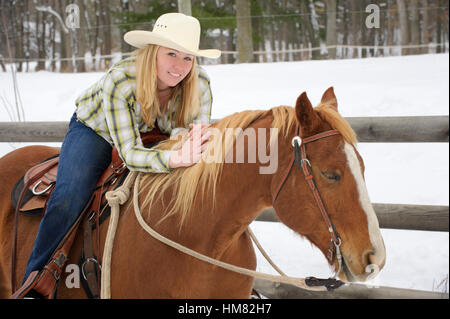 Hübsche blonde Frau, Lächeln, während auf dem Pferderücken, westlichen Freizeitkleidung, weißen Cowboy-Hut, Jeans, kariertes Hemd im Winter tragen. Stockfoto