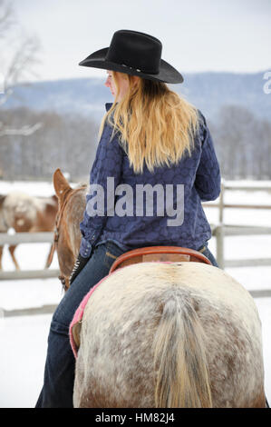 Blonde Frau auf einem grauen Appaloosa Pferd im Winter, junges Cowgirl im westlichen Freizeitkleidung, schwarzer Cowboy-Hut, Jeans Stockfoto