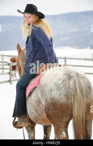 Blonde Frau auf einem grauen Appaloosa Pferd im Winter, junges Cowgirl im westlichen Freizeitkleidung, schwarzer Cowboy-Hut, Jeans Stockfoto