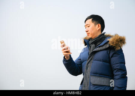 Asiatischer Mann Winterjacke tragen und verwenden ein smart Phone über blauen Himmel Stockfoto