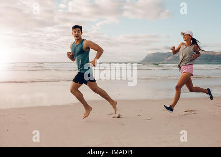 Voller Länge Schuss von Fit junger Mann und Frau am Strand laufen. Glückliches junges Paar Läufer am Ufer Meeres. Stockfoto