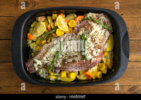 Rohe Lammkeule mit Wurzeln und Zwiebeln zum Braten auf einem rustikalen Holzbrett bereit. Ansicht von oben Stockfoto