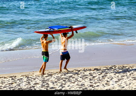 Zwei jungen tragen eine rote Rettung Surfbrett auf dem Kopf entlang der Sand am Kings Beach, Caloundra auf der Sunshine Coast von Queensland, Australien. Stockfoto