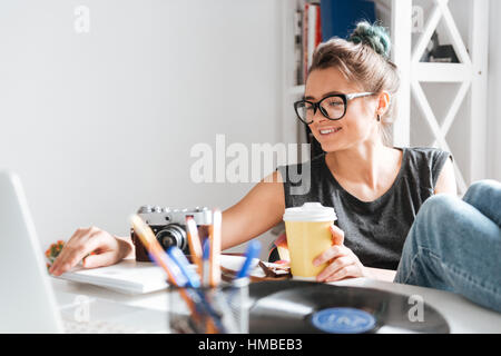 Gerne hübsche junge Frau Fotograf in Gläsern mit Beinen am Tisch sitzen und mit laptop Stockfoto