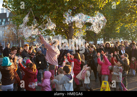 Kleine Kinder und deren Eltern versammeln sich um ein Straßenkünstler hinterließ eine Schneise der riesige Seifenblasen auf der Southbank in London Stockfoto
