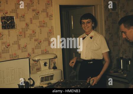 Frau in der 1950er Jahren Küche hält ein Tablett mit Essen und einem Spachtel über den Ofen, eine Kaffeemaschine stehen sehen unten links, einen Mann mit Blick auf das Tablett mit Essen, 1952. Stockfoto