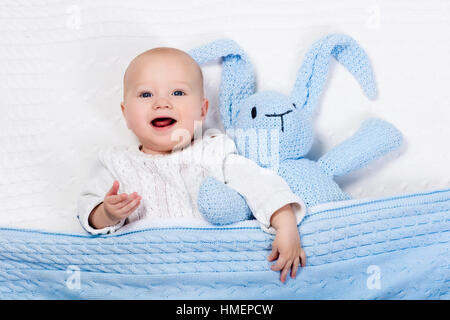 Lustige kleine Baby tragen eine warme Strickjacke, spielen mit Spielzeug Hase auf weißem Kabel stricken Decke im sonnigen Kindergarten Stockfoto