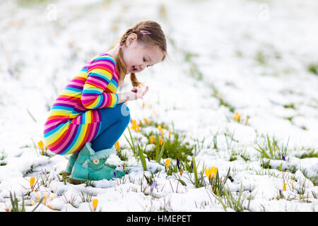 Niedliche kleine Mädchen in buntes Kleid, die ersten Blumen im Frühling Crocus unter Schnee an kalten Tag beobachten. Kind Kommissionierung Garten Blume Stockfoto